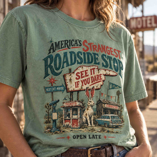 A person wearing a faded sage green graphic t-shirt with a vintage roadside diner and rabbit illustration. The text on the shirt reads 'America's Strangest Roadside Stop' and 'See It If You Dare'.