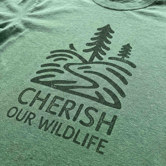 Close-up of a dark green minimal-vintage graphic on a green t-shirt, featuring pine trees, a winding river, and the text "Cherish Our Wildlife".