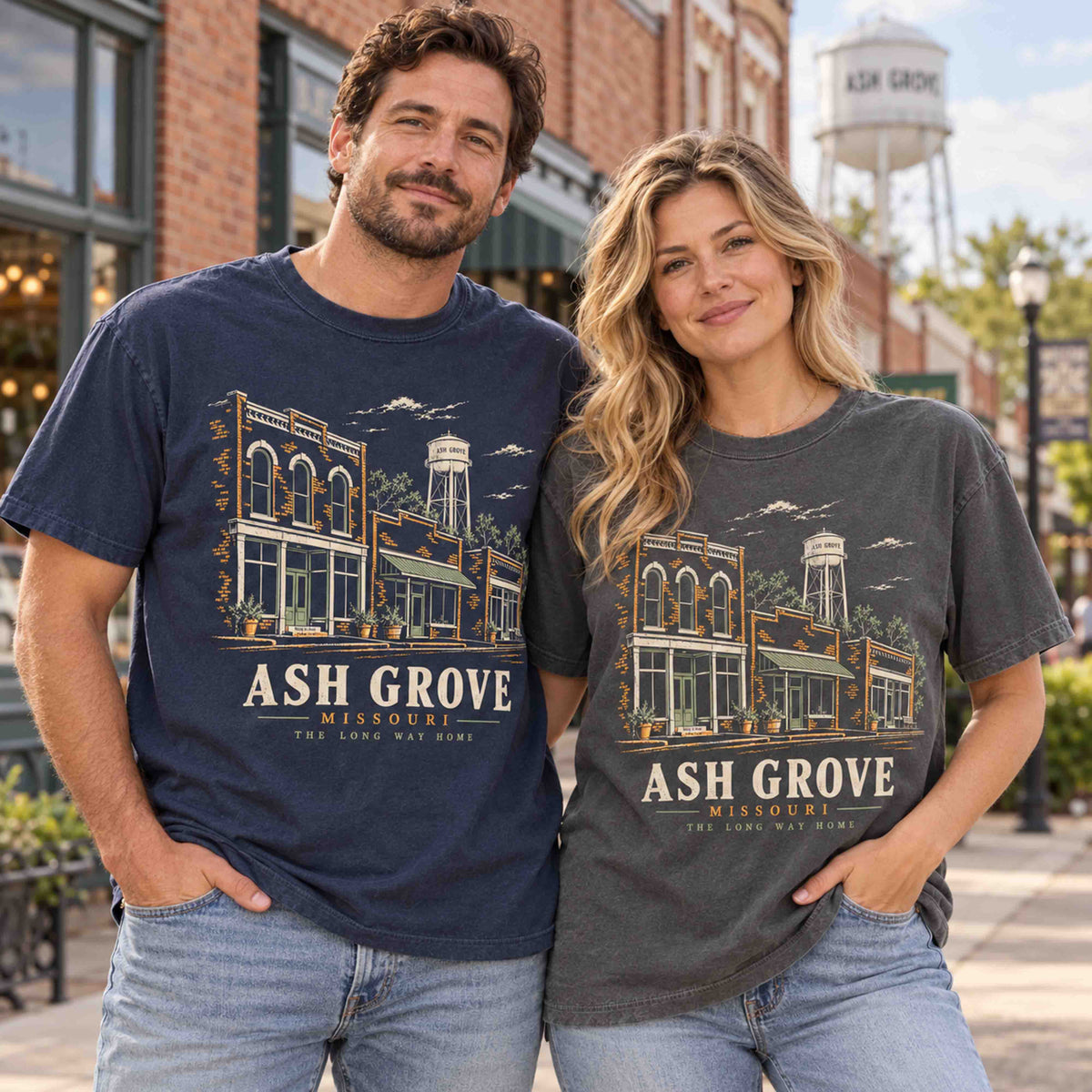 Man and woman wearing Being is Good Ash Grove Missouri graphic T-shirts in Navy and Pepper, featuring storefront and water tower artwork.