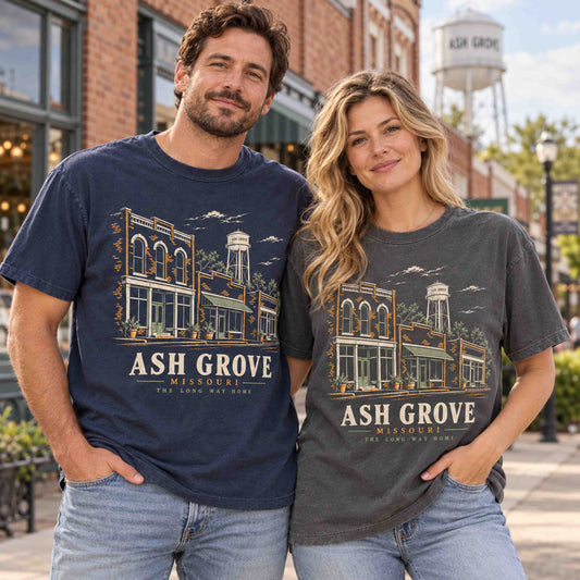 Man and woman wearing Being is Good Ash Grove Missouri graphic T-shirts in Navy and Pepper, featuring storefront and water tower artwork.