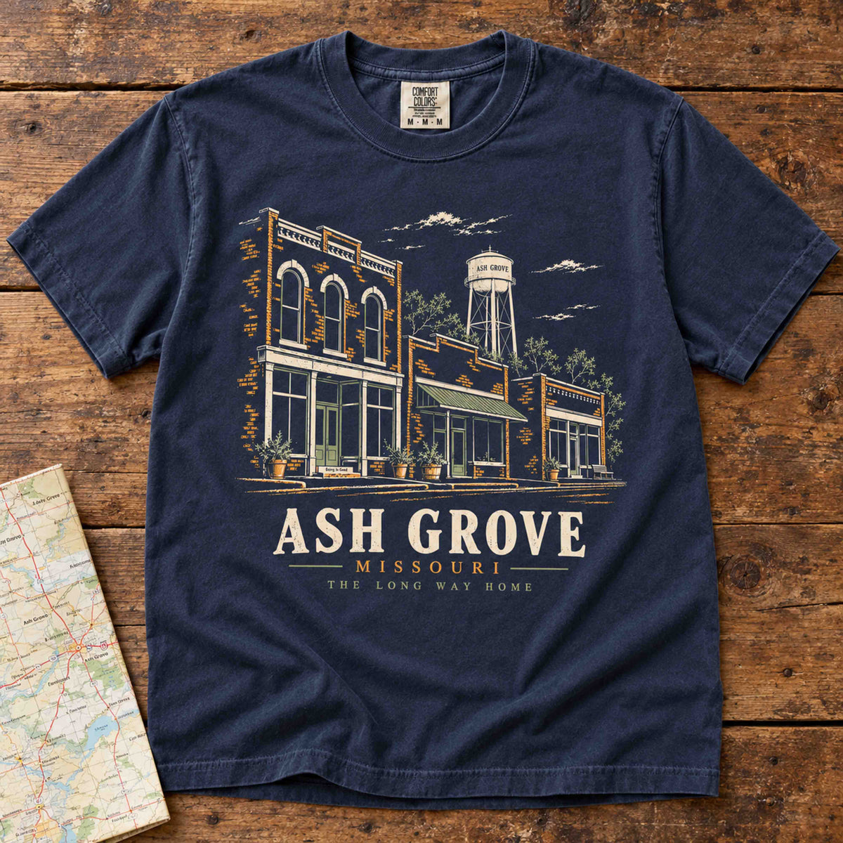 Navy Ash Grove Missouri T-shirt laid flat on a wooden table, showing storefront and water tower artwork.