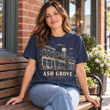 Woman seated on a wooden bench wearing a Navy Ash Grove Missouri graphic T-shirt beside brick storefronts.