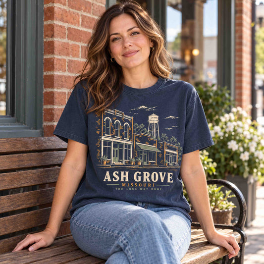 Woman seated on a wooden bench wearing a Navy Ash Grove Missouri graphic T-shirt beside brick storefronts.