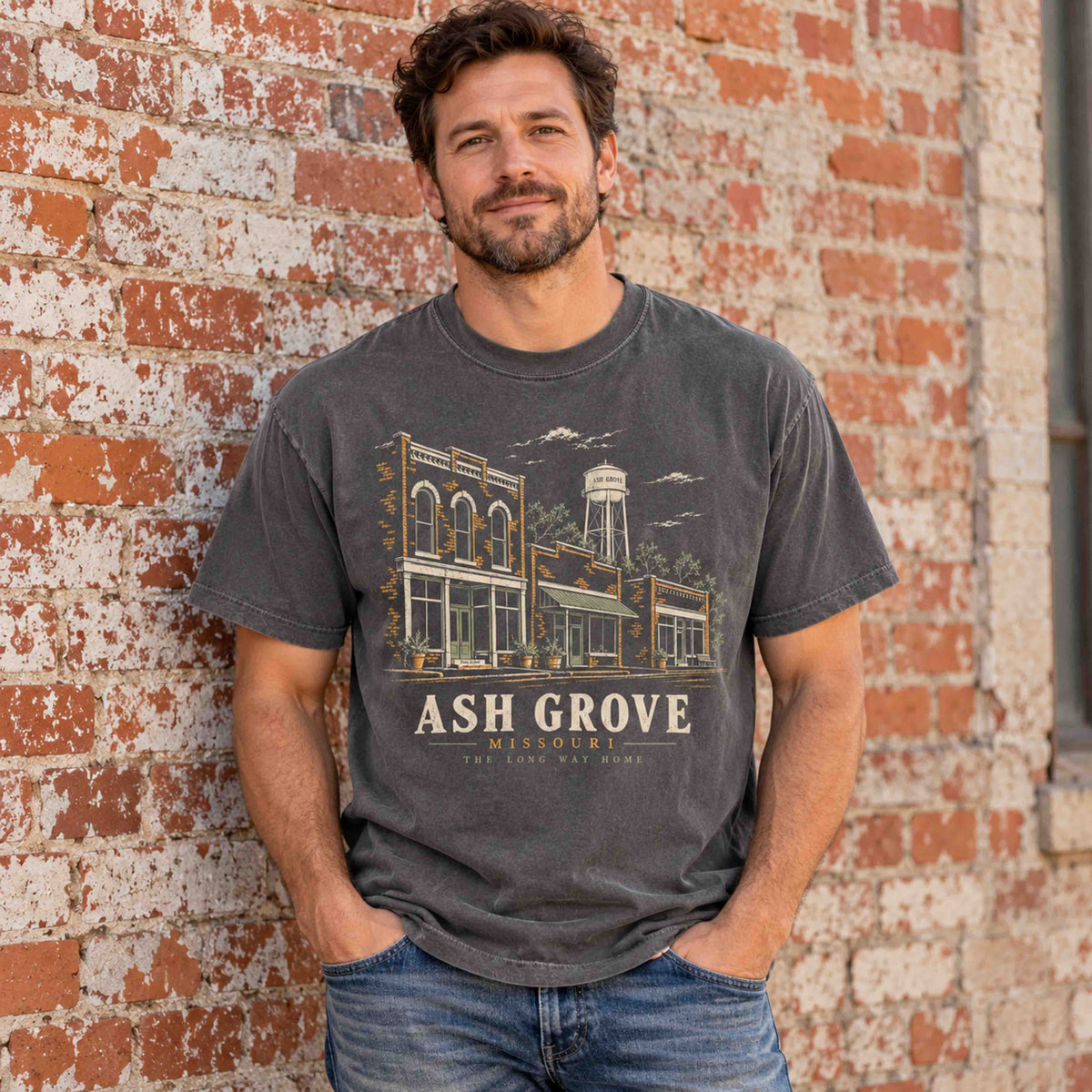 Man wearing a Pepper Ash Grove Missouri graphic T-shirt beside a weathered brick wall.