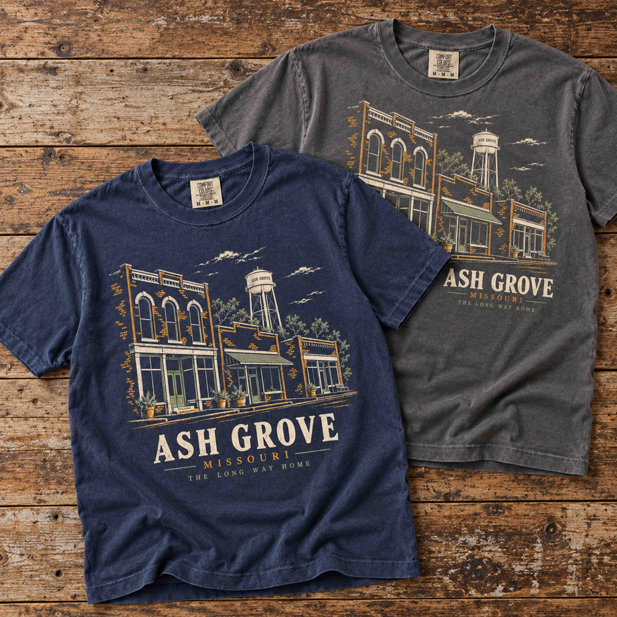 Navy and Pepper Ash Grove Missouri graphic T-shirts overlapping on a rustic wooden table.