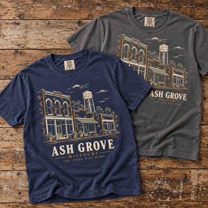 Navy and Pepper Ash Grove Missouri graphic T-shirts overlapping on a rustic wooden table.