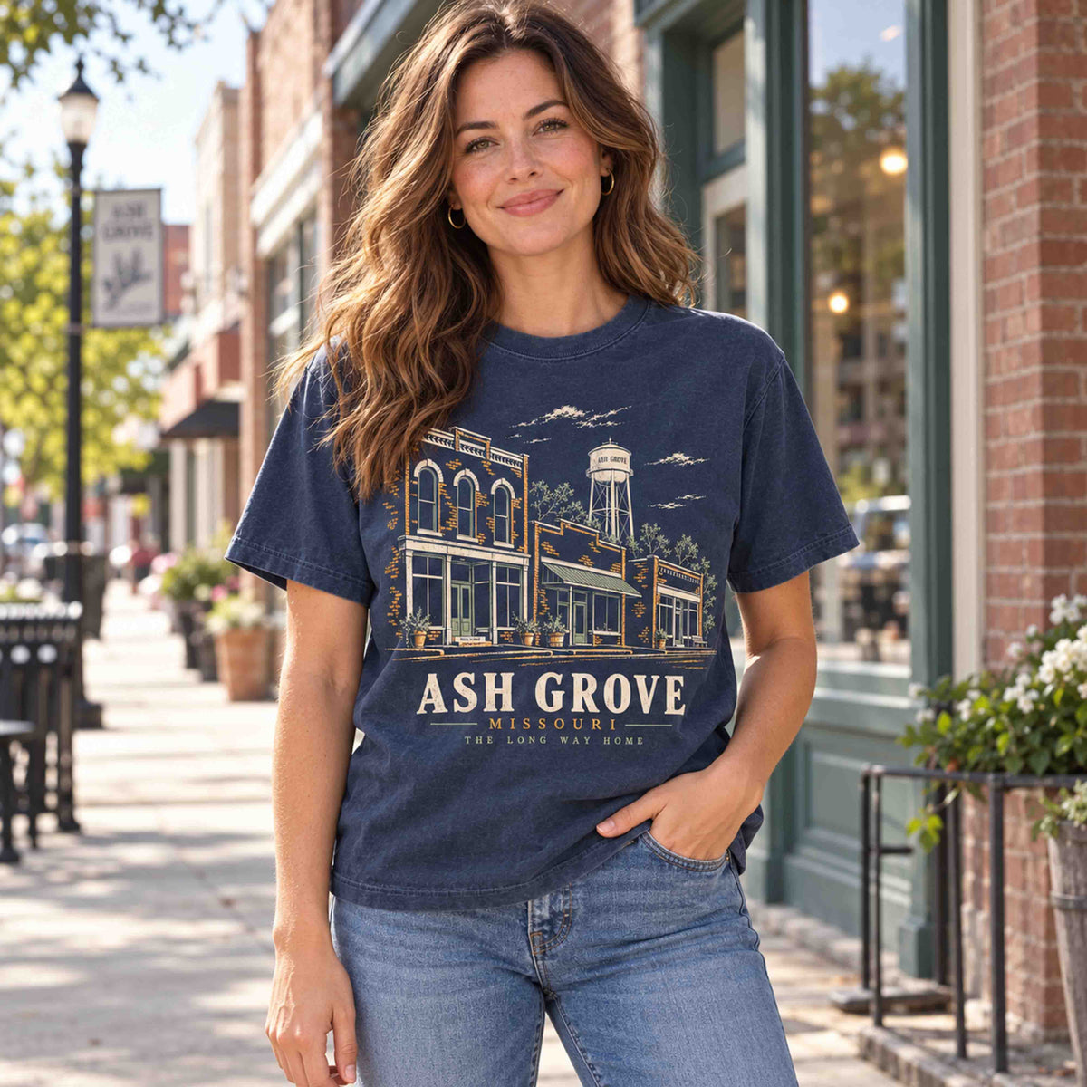 Woman wearing a Navy Ash Grove Missouri graphic T-shirt on a sidewalk lined with brick storefronts.