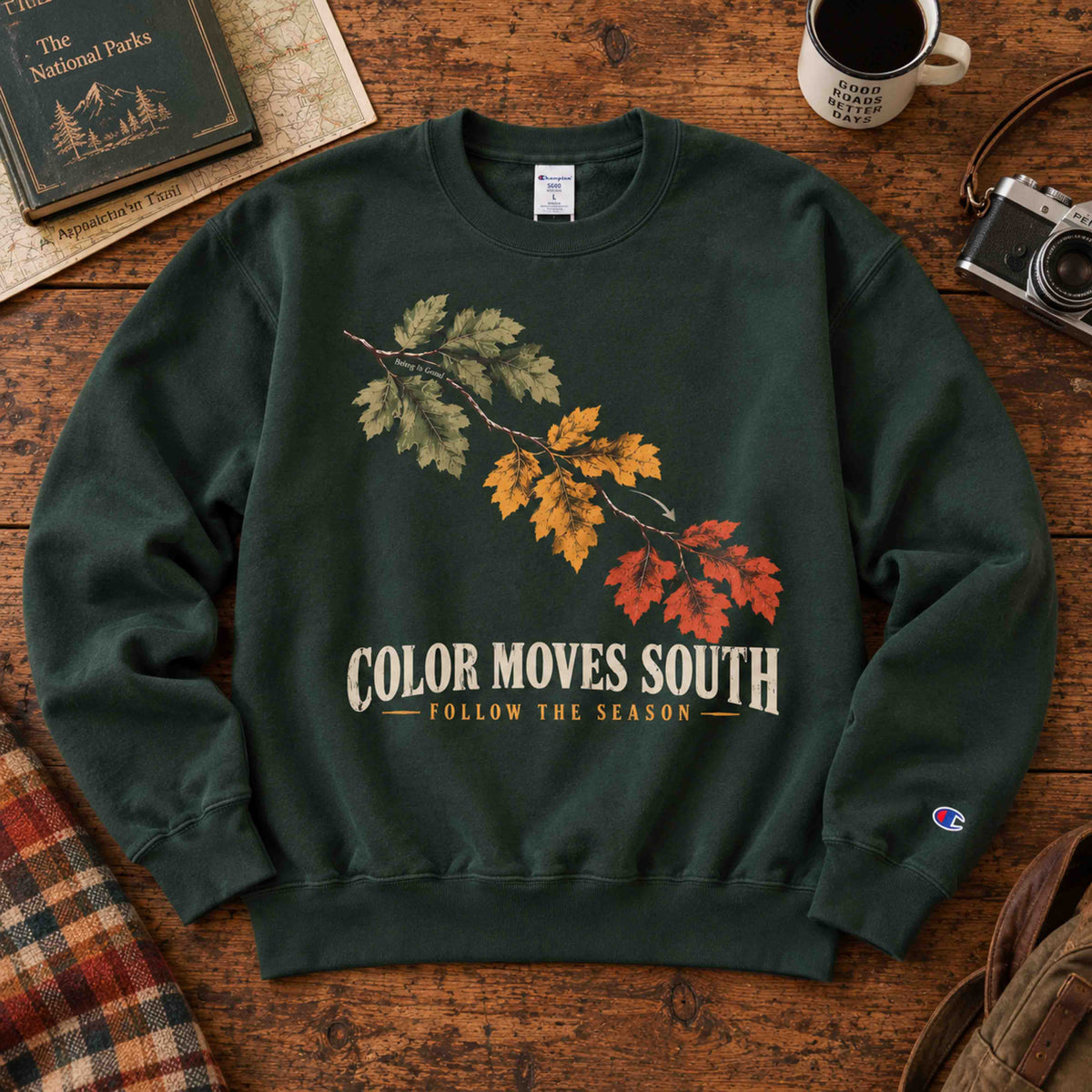 Being is Good Color Moves South sweatshirt in Dark Green, laid flat on a wooden table with autumn leaves artwork.