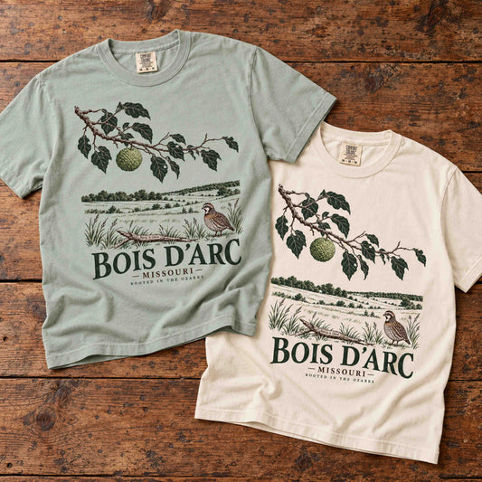 Bay and Ivory Bois D'Arc Missouri graphic T-shirts laid together on a rustic wooden table.