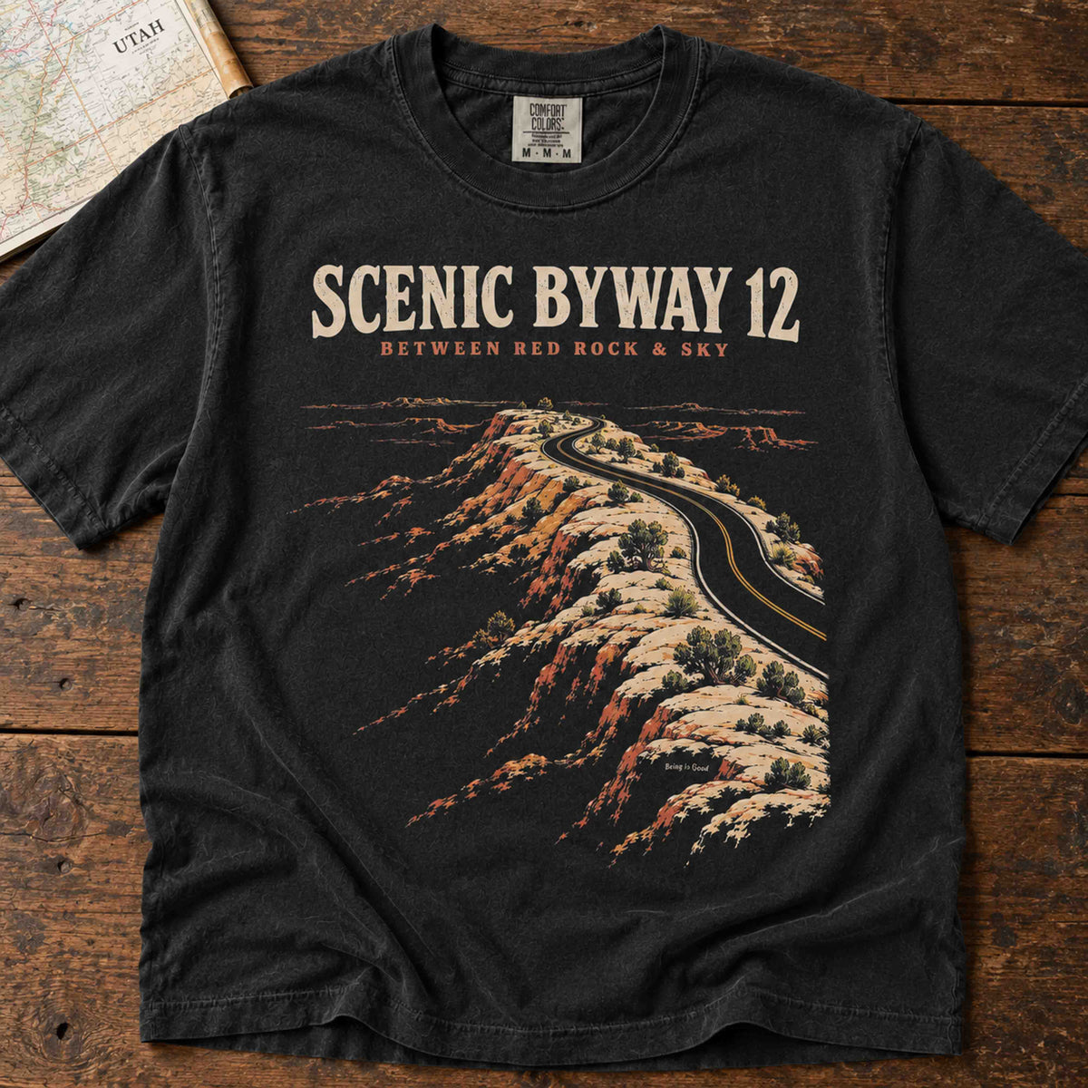 Black Scenic Byway 12 graphic T-shirt laid flat, featuring a winding desert highway and Between Red Rock & Sky lettering.