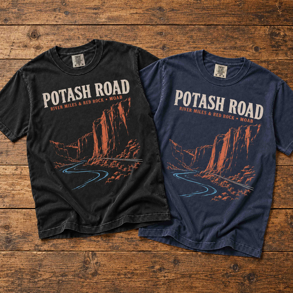 Black and Navy Being is Good Potash Road graphic T-shirts laid side by side on a wooden surface.