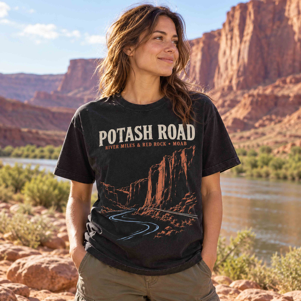 Woman wearing a Black Potash Road Moab T-shirt with cream lettering, rust-colored canyon cliffs and blue river artwork.