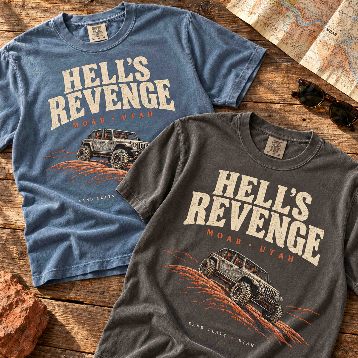 Blue Jean and Pepper Hell’s Revenge Moab graphic t-shirts arranged diagonally on a vintage wooden table.