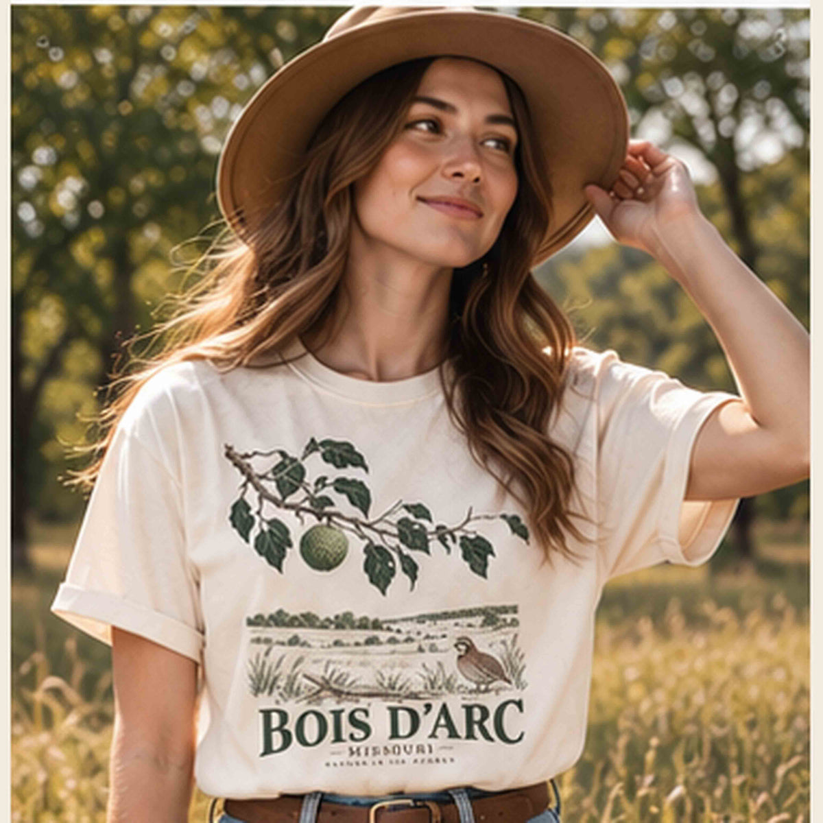 Woman wearing an Ivory Bois D'Arc Missouri graphic T-shirt and a wide-brim hat in a countryside setting.