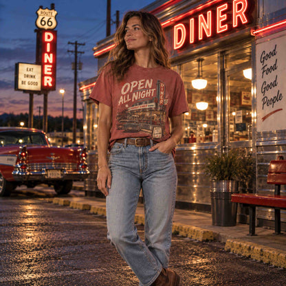 A woman stands on a diner street corner at dusk wearing a faded mauve graphic T-shirt with the words 'OPEN ALL NIGHT' and light-wash jeans. The diner sign glows in the background, along with a red classic car and wet pavement.