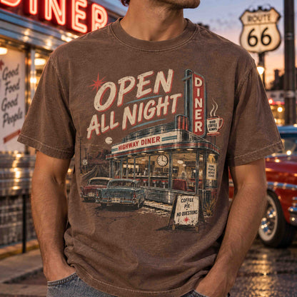 A man wearing a brown vintage-style graphic t-shirt featuring a retro diner scene with the text 'OPEN ALL NIGHT'. The background shows a blurred nighttime street scene with a Route 66 sign and a diner.
