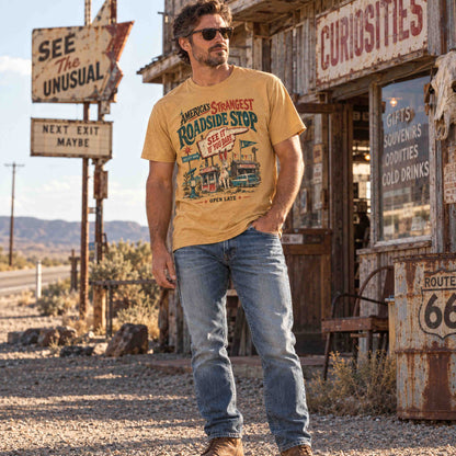A man wearing a tan graphic t-shirt and blue jeans stands in a rustic, desert-themed western town street scene. The t-shirt features a vintage-style roadtrip graphic with the text 'America's Shortest Roadside Stop See The Stranger'.