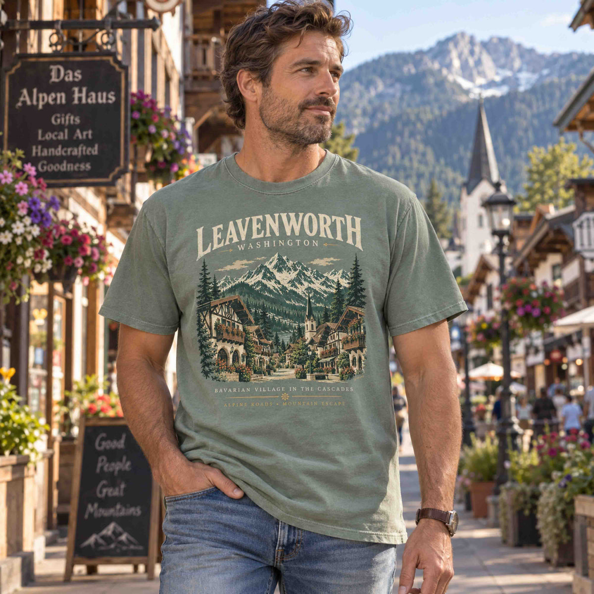 Man wearing a Moss green Leavenworth Washington graphic tee on a Bavarian-style village street with mountains behind him.