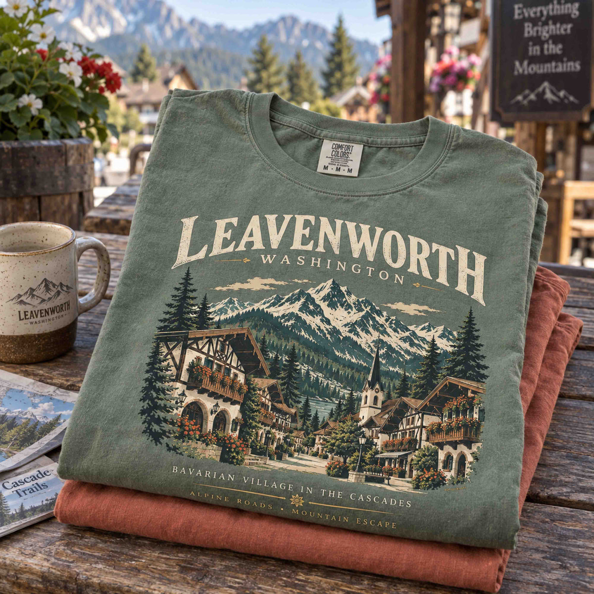 Folded Moss green Leavenworth Washington graphic tee stacked over a Yam tee, showing Bavarian village and mountain artwork.