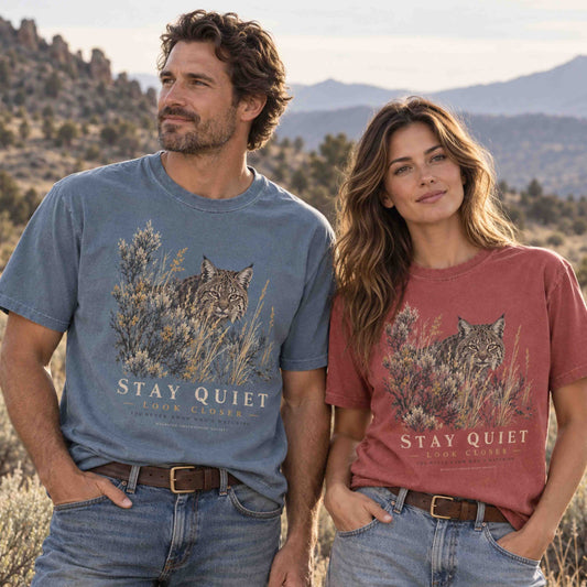 Couple wearing Stay Quiet Look Closer bobcat wildlife graphic tees in Blue Jean and Crimson in a desert mountain landscape