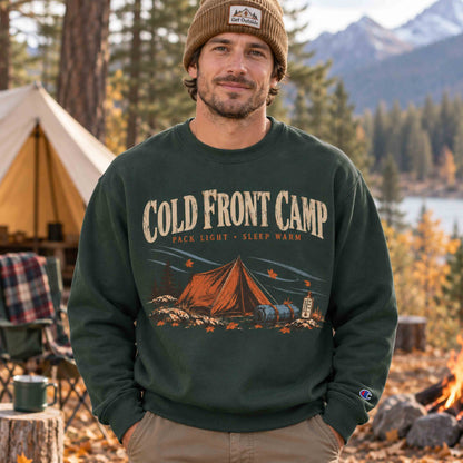 Dark Green Cold Front Camp graphic sweatshirt worn by man sitting beside a campfire at an autumn campsite