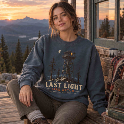 Woman wearing a Pepper Last Light Fire Watch graphic sweatshirt at sunset near a mountain lookout tower