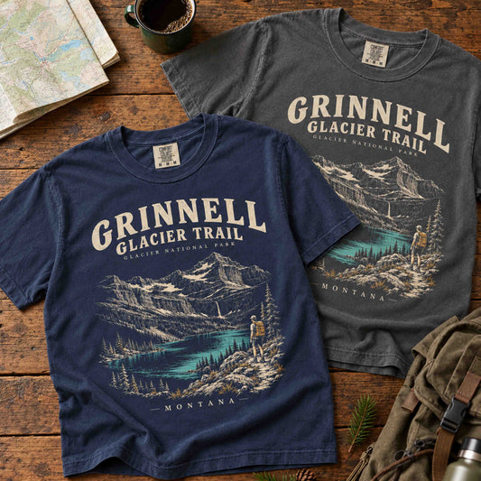 Navy and Pepper Grinnell Glacier Trail graphic T-shirts arranged diagonally on a wooden table beside a map and hiking gear.