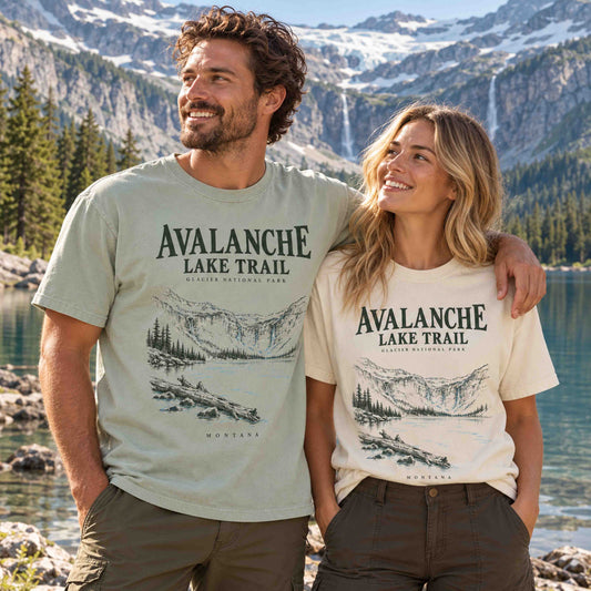 Man and woman wearing Being is Good Avalanche Lake Trail Glacier National Park T-shirts in Bay and Ivory beside a mountain lake.