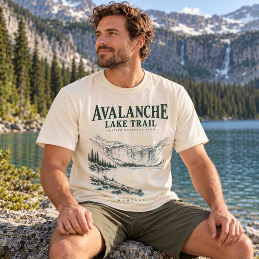 Man seated on a rock wearing an Ivory Avalanche Lake Trail Glacier National Park graphic T-shirt beside a lake.