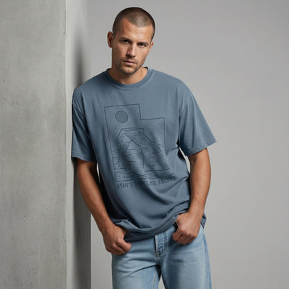 Man looking down with his thumbs in his pockets, wearing a vintage-style blue Utah desert t-shirt.