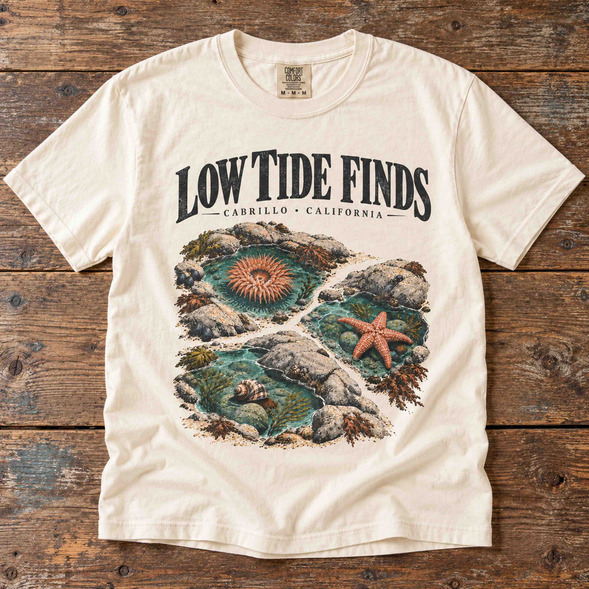 Overhead view of the Ivory Low Tide Finds Cabrillo graphic tee on a wooden table, showing tide pools and marine life.