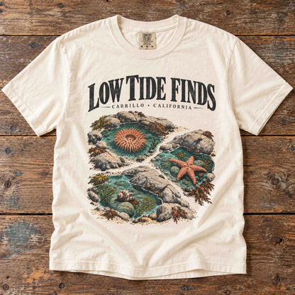 Overhead view of the Ivory Low Tide Finds Cabrillo graphic tee on a wooden table, showing tide pools and marine life.