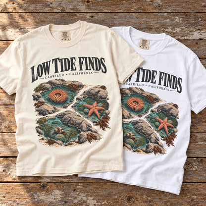 Ivory and White Low Tide Finds Cabrillo graphic tees layered on a wooden table with tide pool artwork visible.