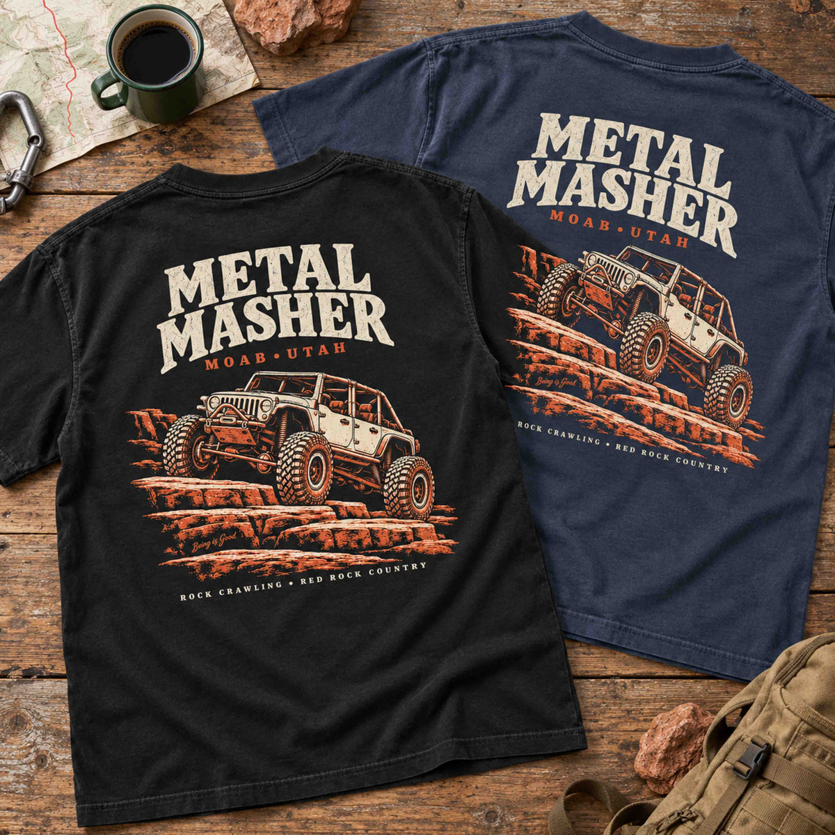 Black and Navy Metal Masher tees on a wood table, both showing large rock-crawling back graphics.