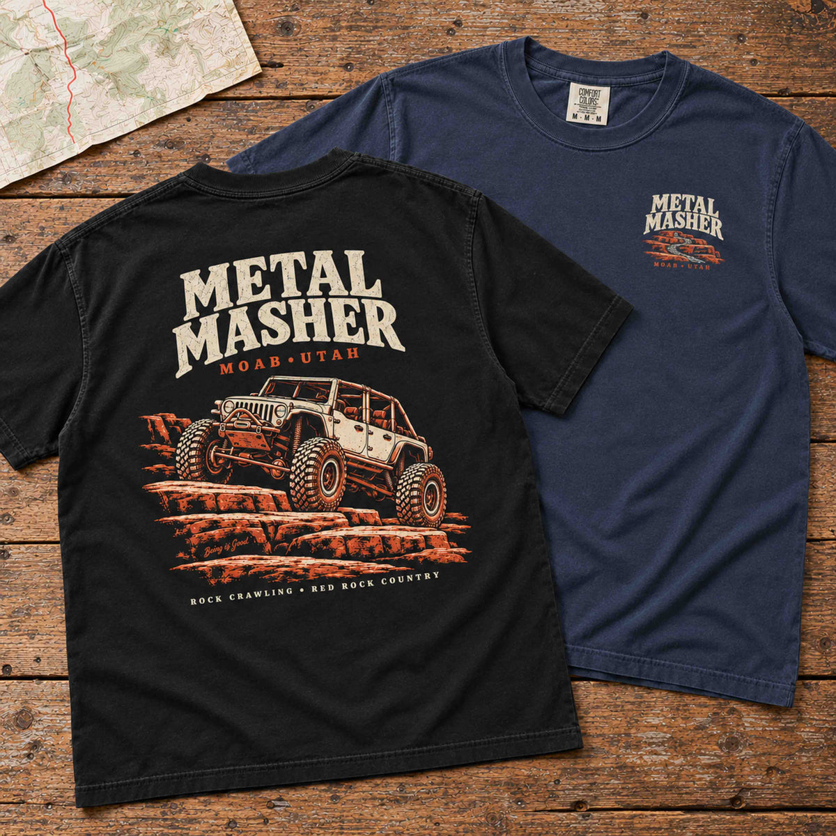 Black Metal Masher tee showing the back graphic beside a Navy tee showing its small left-chest print on a wood table.