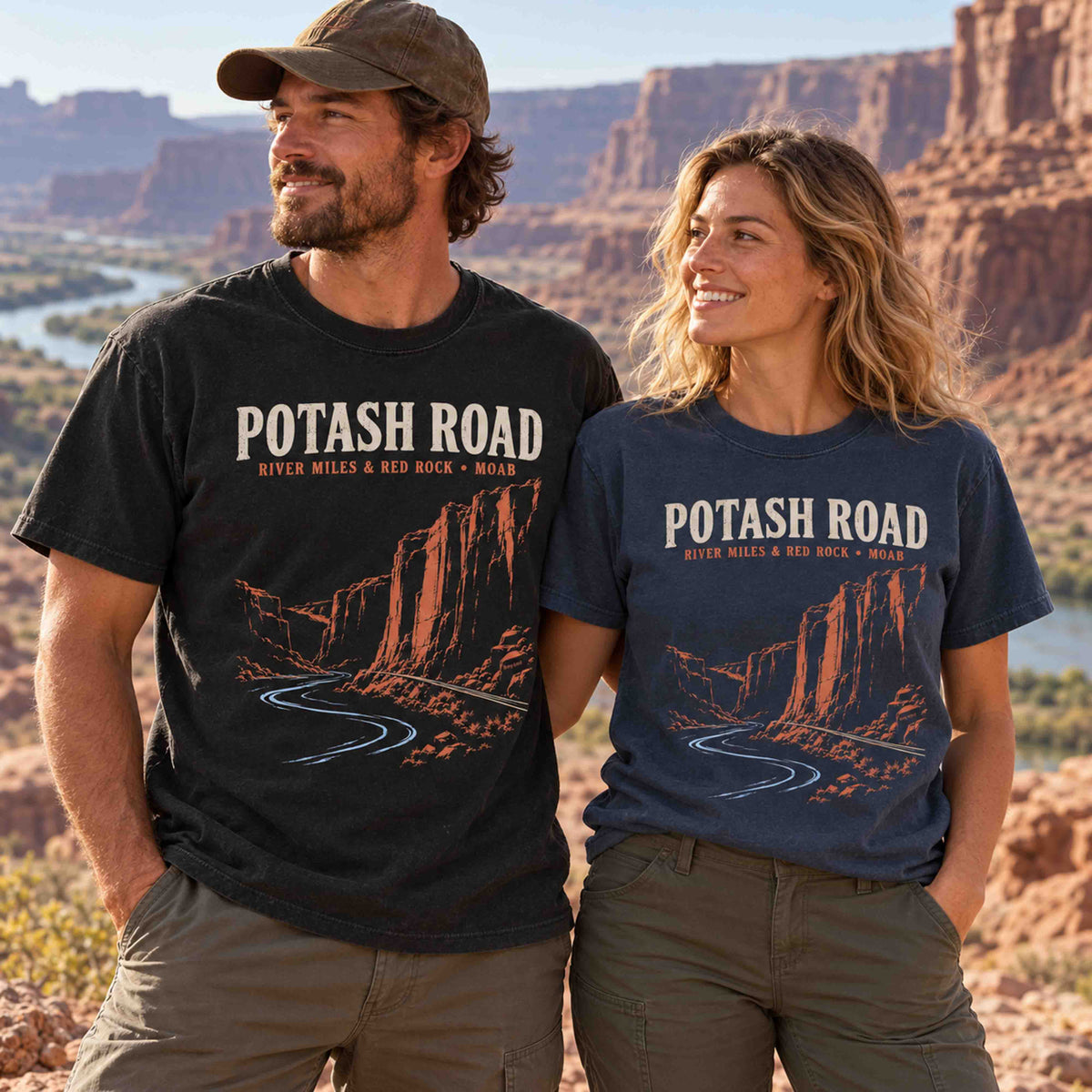 Man and woman wearing Being is Good Potash Road Moab graphic T-shirts in Black and Navy beside red rock cliffs and a river.