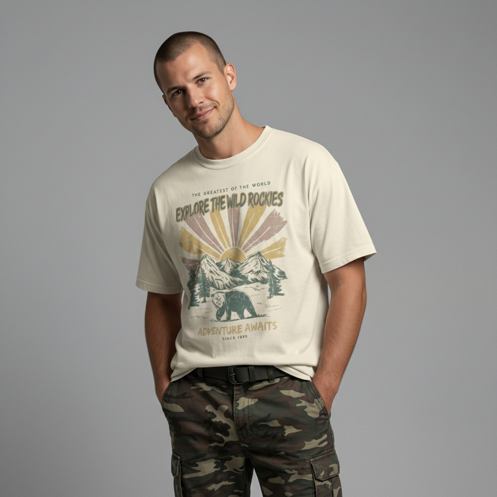 Man wearing a cream-colored Being is Good t-shirt with a vintage "Explore the Wild Rockies" bear and mountain graphic, paired with camo cargo pants.