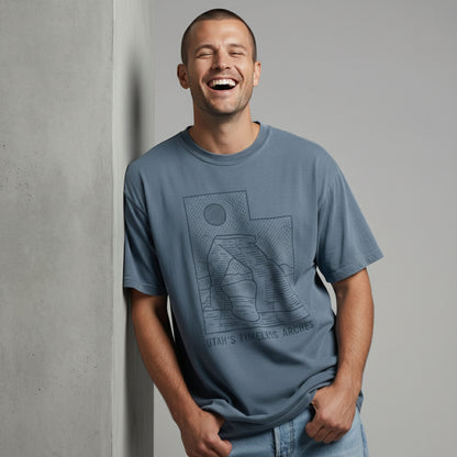 Man laughing and leaning against a wall, wearing a relaxed-fit dusty blue t-shirt with a minimal Utah arches graphic.