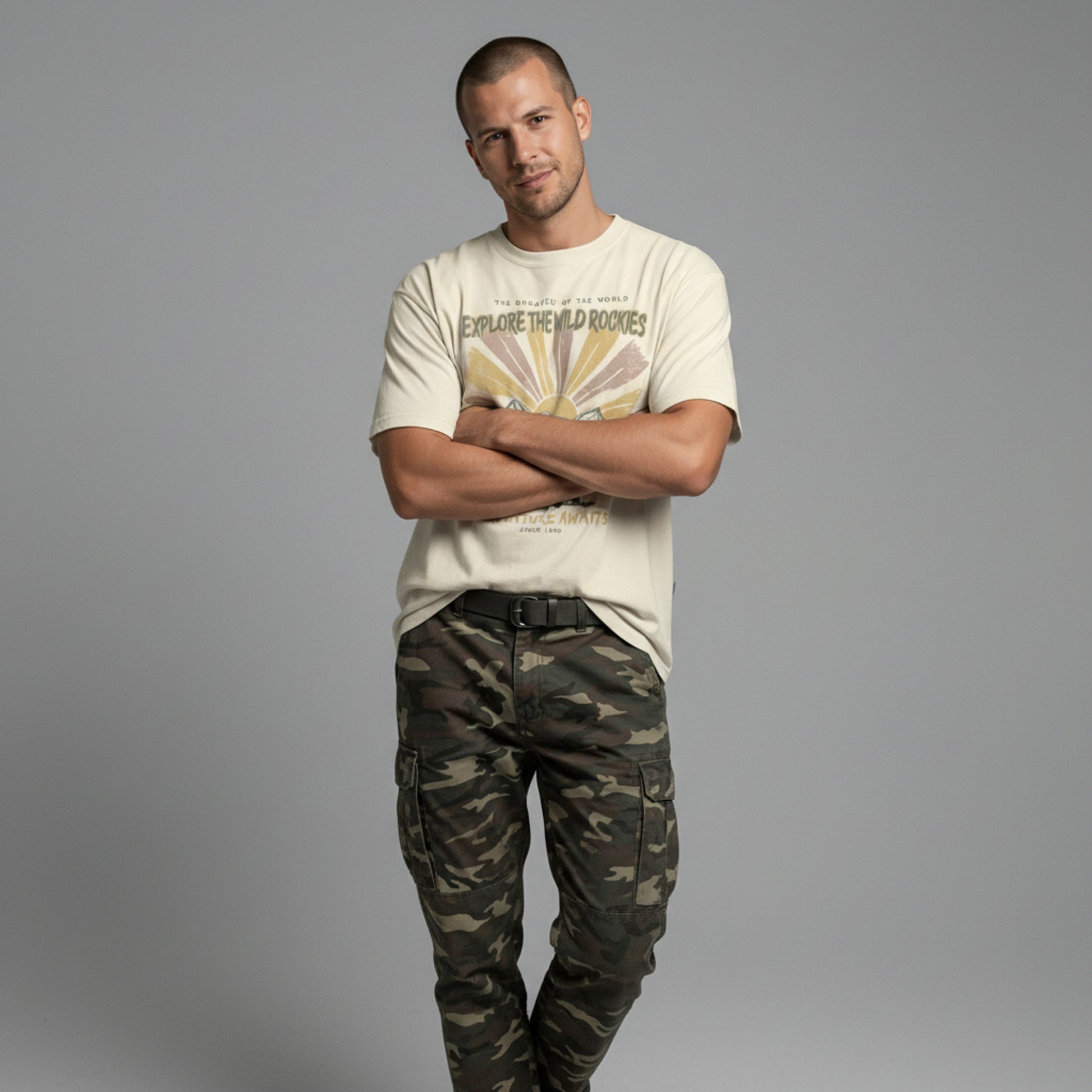 Front view of a man with arms crossed, wearing a cream vintage graphic t-shirt and camouflage pants against a solid grey background.