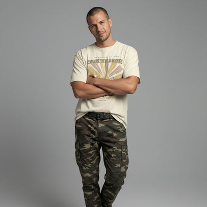 Front view of a man with arms crossed, wearing a cream vintage graphic t-shirt and camouflage pants against a solid grey background.