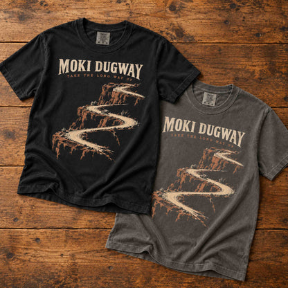Black and Pepper Moki Dugway road trip T-shirts laid diagonally on a rustic wood surface.