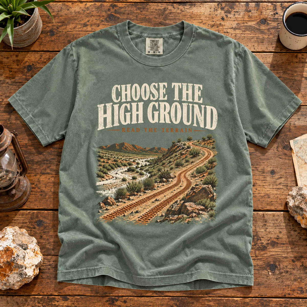 Overhead view of the Moss Choose the High Ground graphic tee on a wooden table with desert trail artwork.