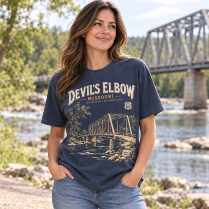 Woman wearing a Navy Devil's Elbow Missouri Route 66 graphic T-shirt beside a river with a steel truss bridge in the background.