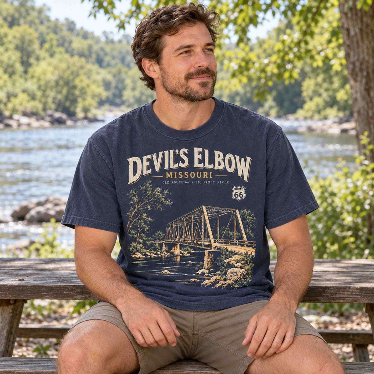 Man seated beside a river wearing a Navy Devil's Elbow Missouri Route 66 graphic T-shirt by Being is Good.