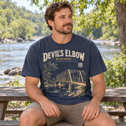 Man seated beside a river wearing a Navy Devil's Elbow Missouri Route 66 graphic T-shirt by Being is Good.