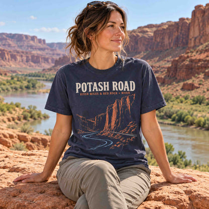 Woman seated on a red rock wearing a Navy Potash Road Moab graphic T-shirt, with a river and canyon in the background.