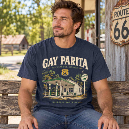 Man seated on a rustic bench wearing a Navy Gay Parita Route 66 graphic T-shirt beside a Route 66 sign.
