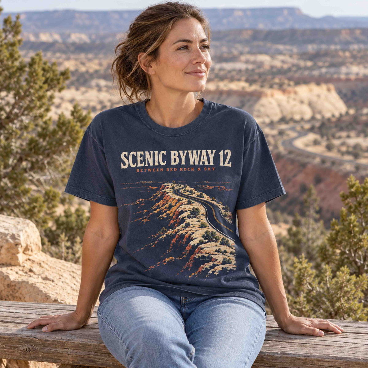 Woman seated outdoors wearing a navy Scenic Byway 12 graphic T-shirt with Utah red rock highway artwork.
