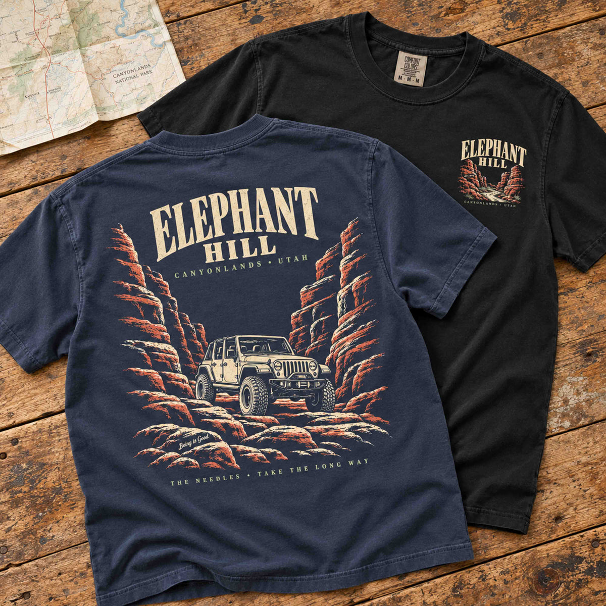 Navy Elephant Hill tee showing its back graphic beside a Black tee showing the small front print on a wood table.