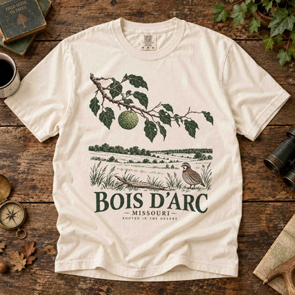 Ivory Bois D'Arc Missouri T-shirt laid flat on a wooden table, showing Osage orange branch and bobwhite quail artwork.